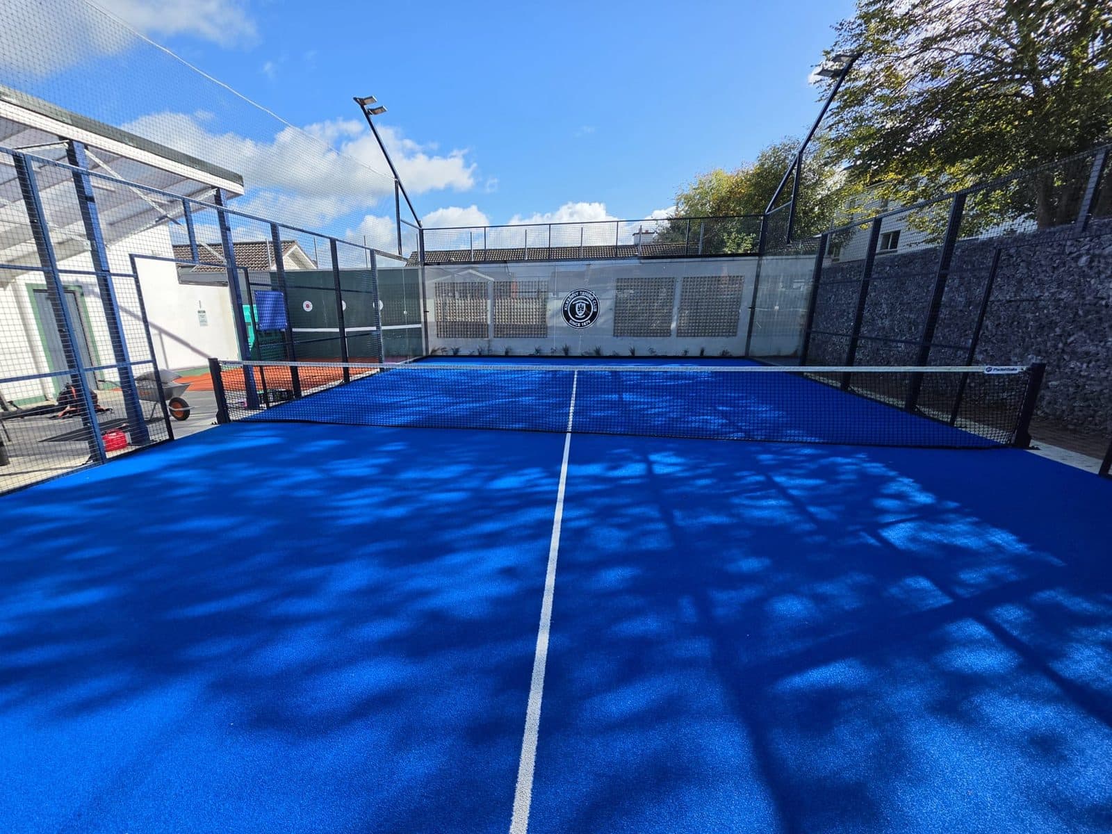 A blue synthetic padel court with white lines, a net and glass walls, in sunshine