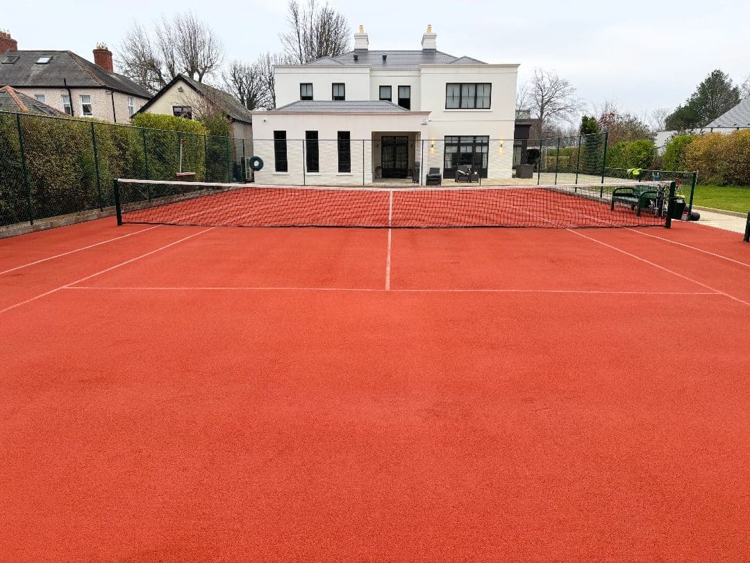 Completed red artificial clay tennis court with net rigged and crisp white line markings, behind a private residence
