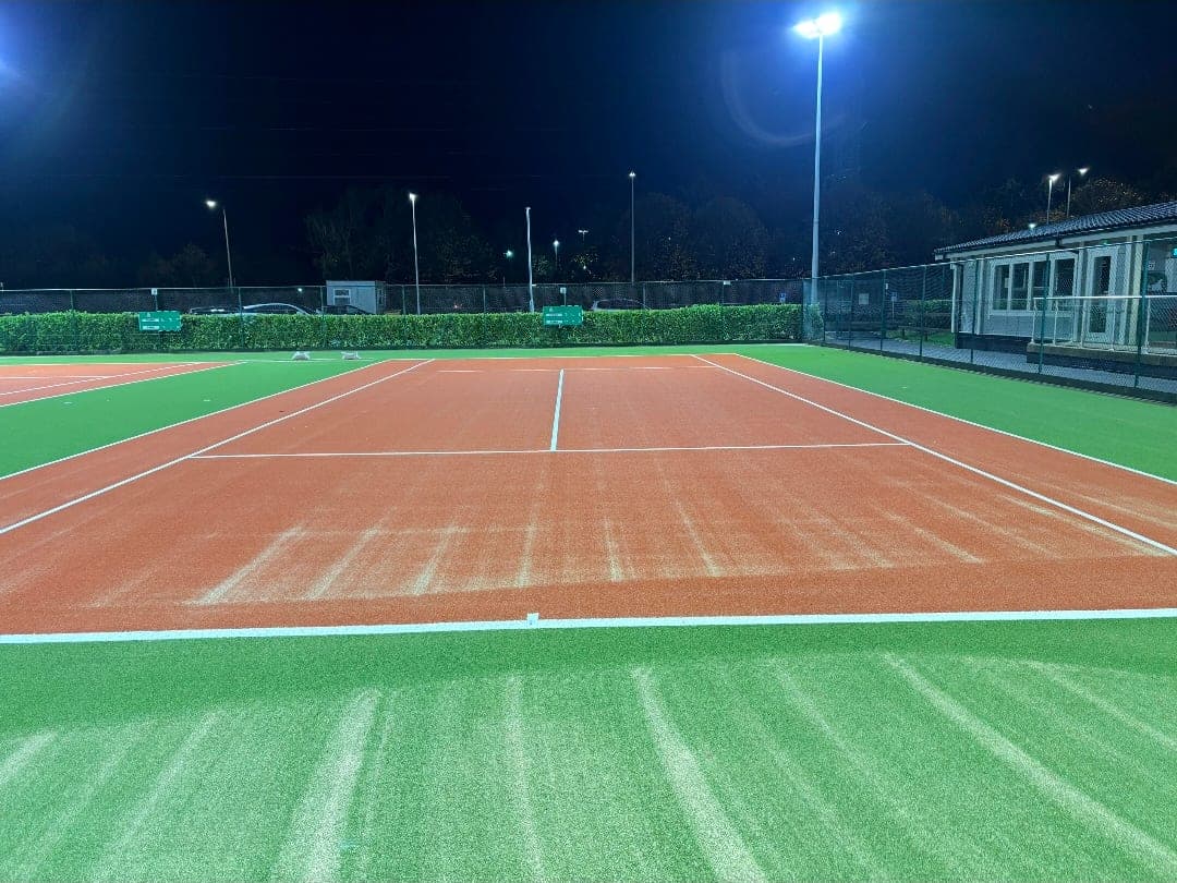 Synthetic tennis court with red playing area and green run-off, lit by LED floodlights at night