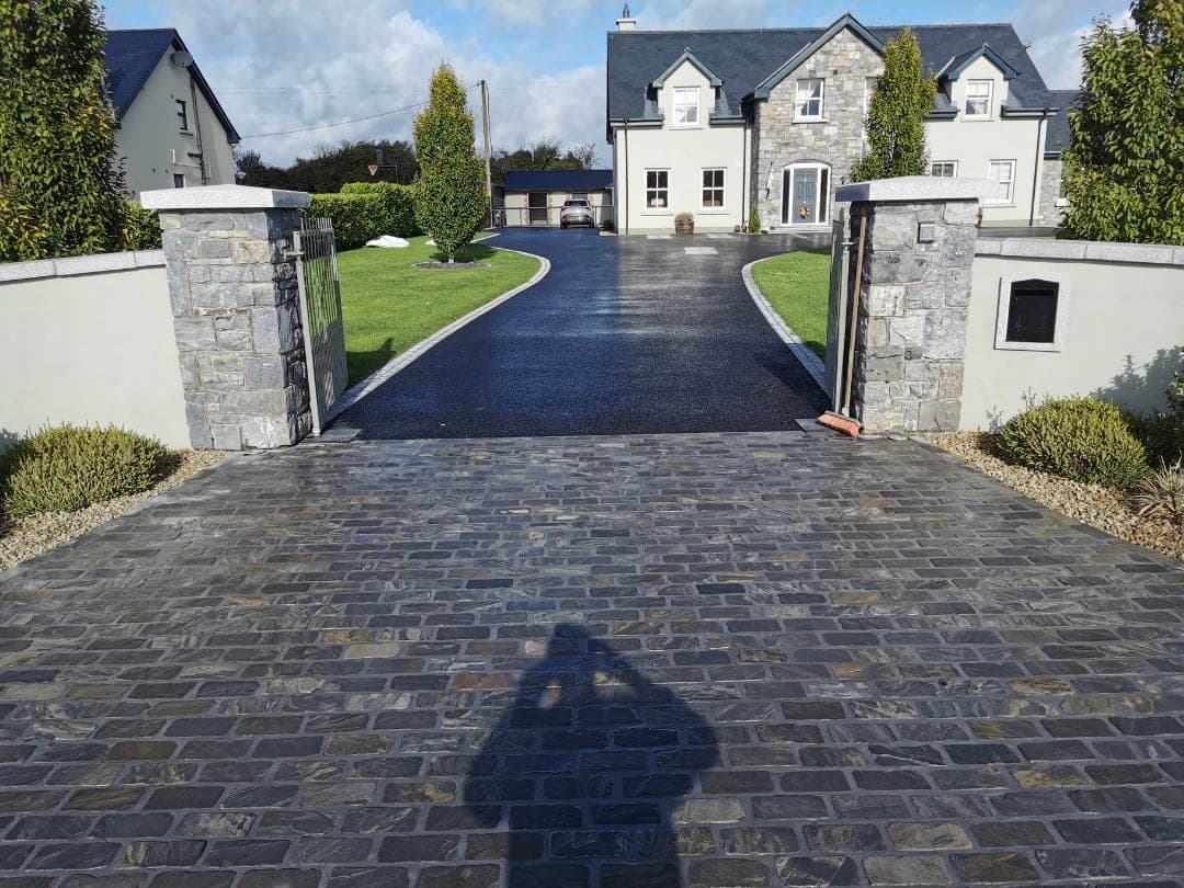 Stone-sett entrance apron between granite piers leading to a tarmac driveway and house