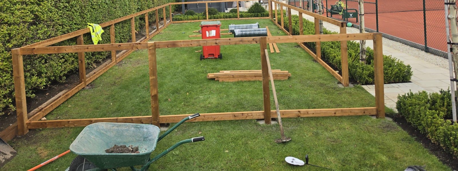 Timber posts and rails part built around a lawn, with timber lengths and rolls of mesh on the grass